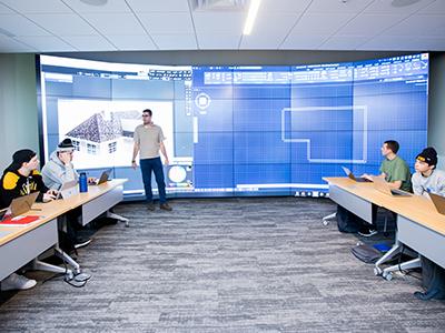 A professor stands in front of 24 television screens with a blueprint blown up on it in a classroom